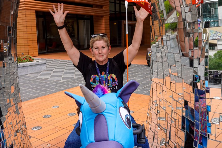 Person on a mobility scooter dressed as a blue unicorn under a mirrored arch, raising hands.