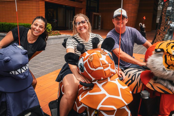 Three people smiling on animal-themed scooters outside a building.