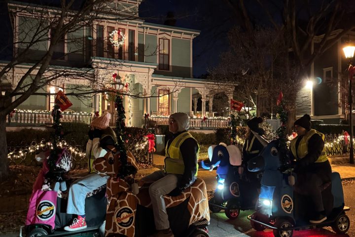 People riding animal-themed scooters with lights in front of a decorated house at night.