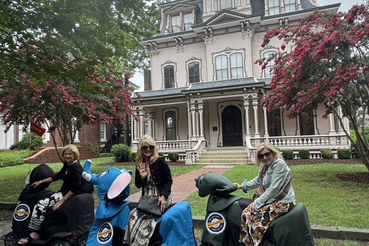 People in costumes posing on small carts in front of a Victorian-style building.