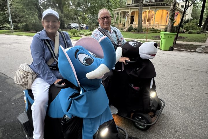 Group in costumes with a blue character on a scooter, on a street with trees and a house in the background.