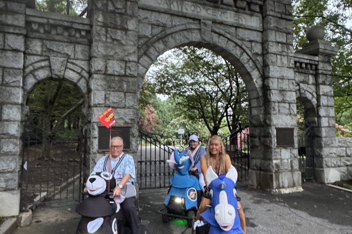 Three people on scooters under stone archway on a paved path.