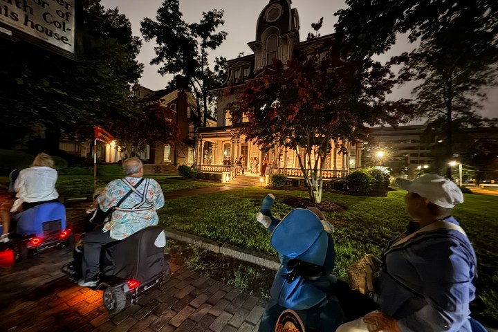 People on mobility scooters outside a lit-up, historic house at night.