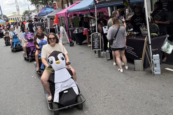 Man rides small penguin-themed cart at outdoor street fair with tents and people.