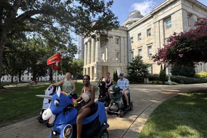 People on mobility scooters outside a large building with flags and trees.