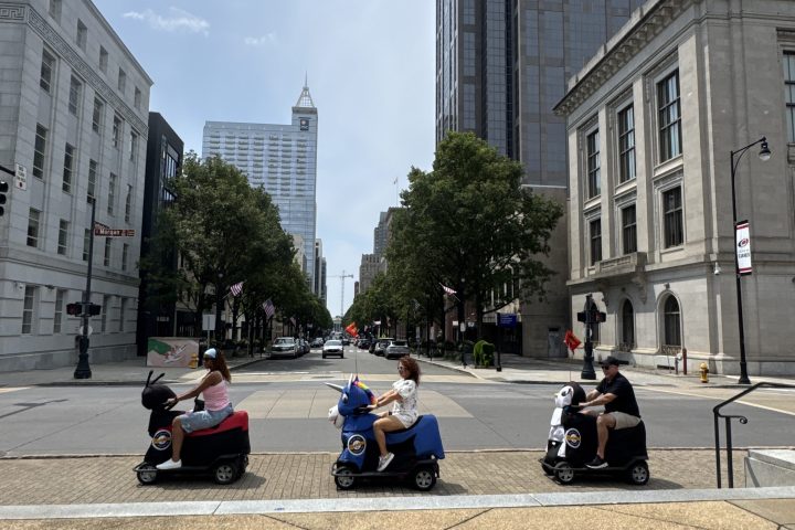People on scooters in an urban area with tall buildings and trees.