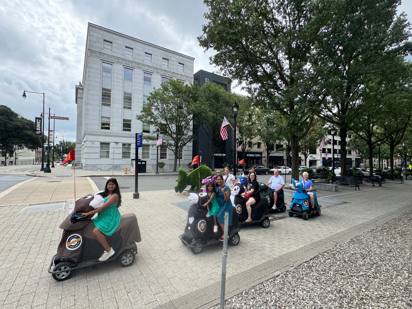 Group riding mobility scooters on a city sidewalk near trees and buildings.