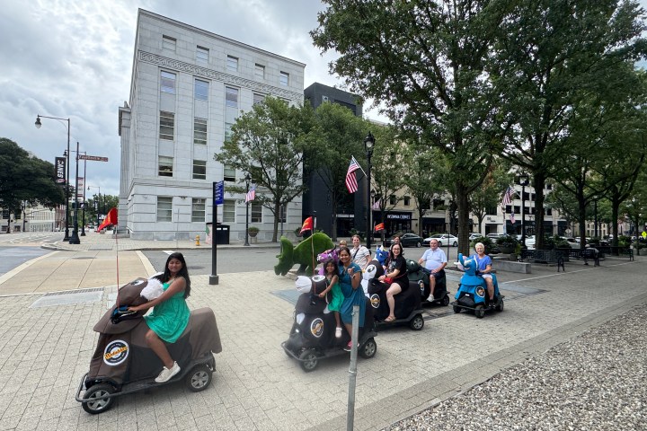 Group riding mobility scooters on a city sidewalk near trees and buildings.