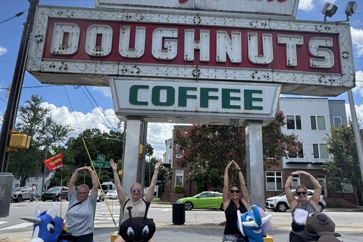 Group posing playfully under a vintage Krispy Kreme doughnuts sign on a sunny day.