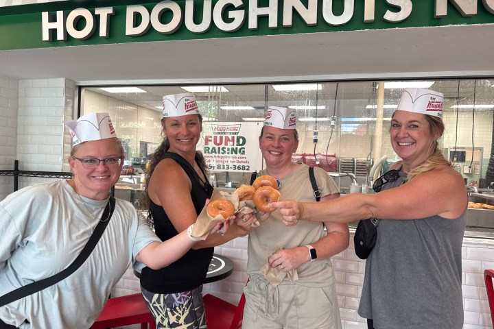 Four people smiling and holding doughnuts in front of a Krispy Kreme shop.