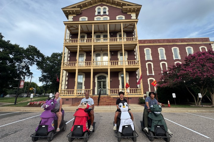 Four people on animal-shaped scooters in front of a historic brick building.