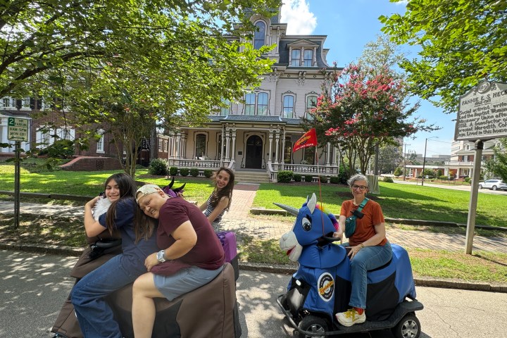 Four people on scooters, one shaped like a donkey, in front of a Victorian house.