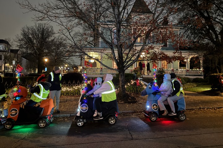People on decorated scooters with lights in front of a large, lit-up house at dusk.