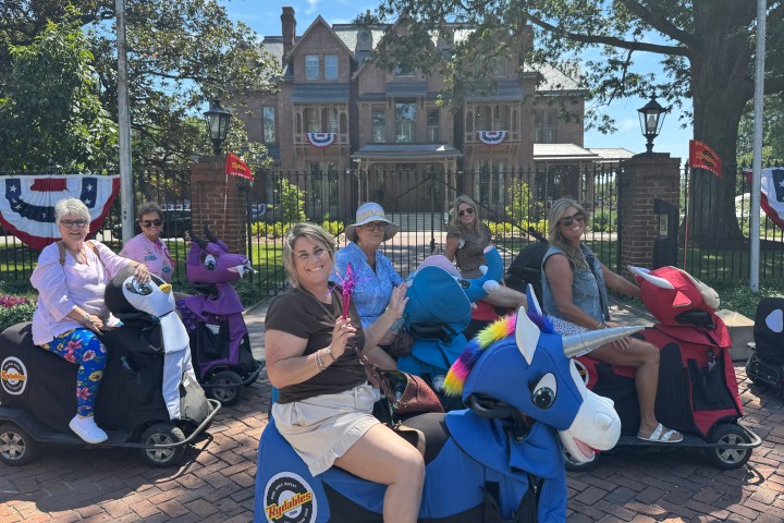 Adults ride motorized animal scooters outside a large house with festive decor.