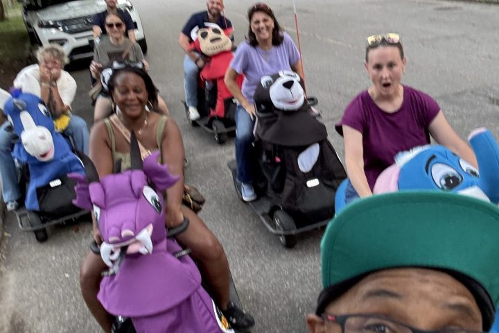 Smiling group in costume parade on scooters or strollers.