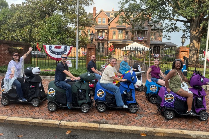 Group of people on motor scooters with animal covers in front of a decorated house.