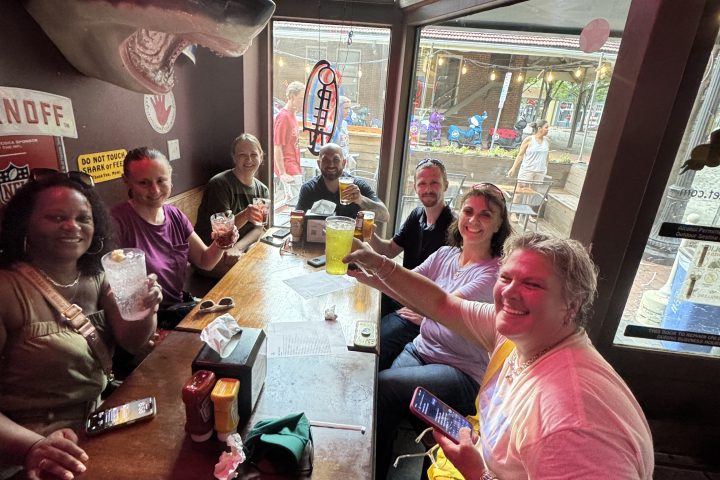 Group of people sitting around a table in a bar, smiling and holding drinks, with a TV on in the background.