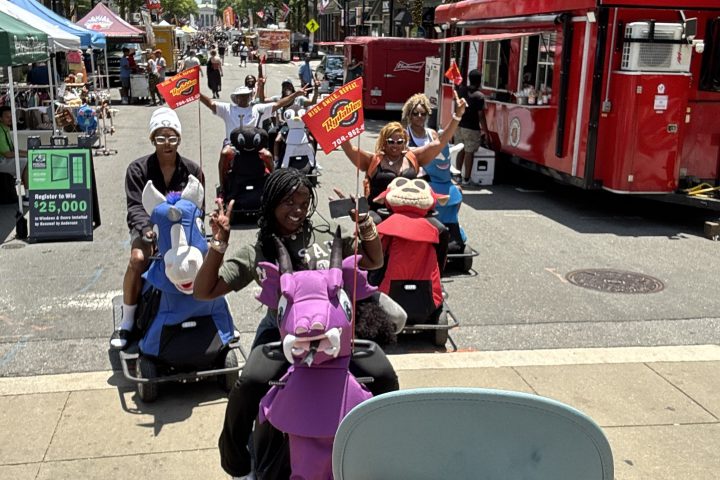 Smiling group in wheelchairs on city street with red bus and trees.