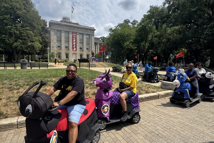 People in animal-themed scooters on a sidewalk, with trees and a building in the background.