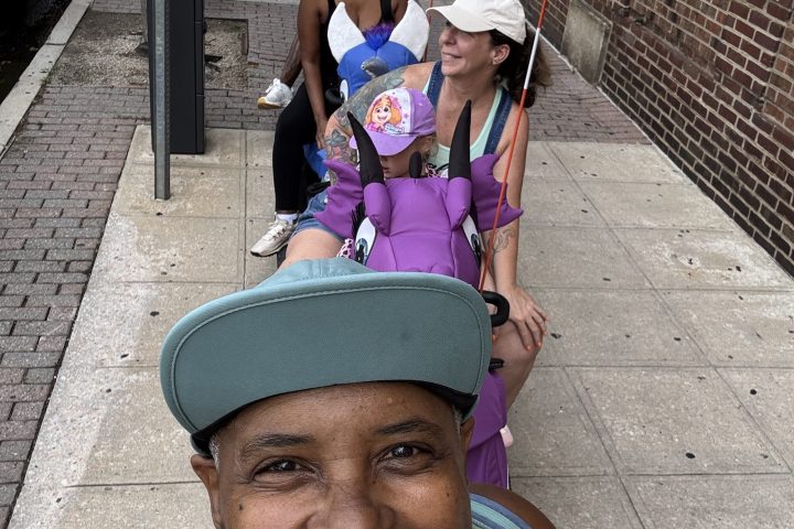 Group selfie of four people smiling, holding festival flags on a city sidewalk.