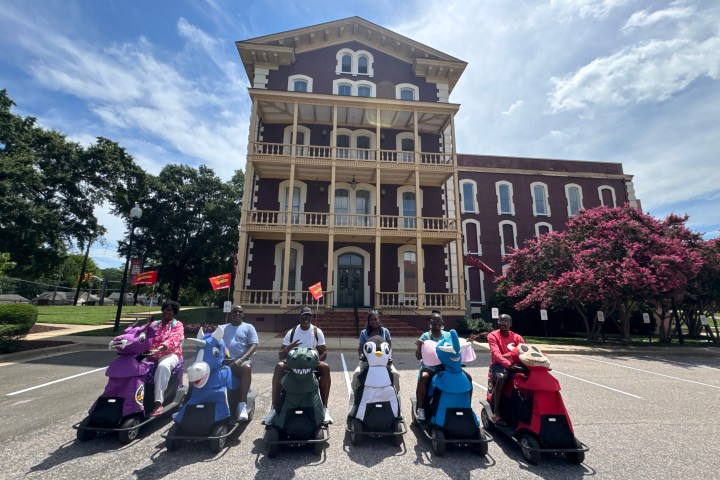 Seven people on scooters in front of historic three-story building with trees and clear sky.