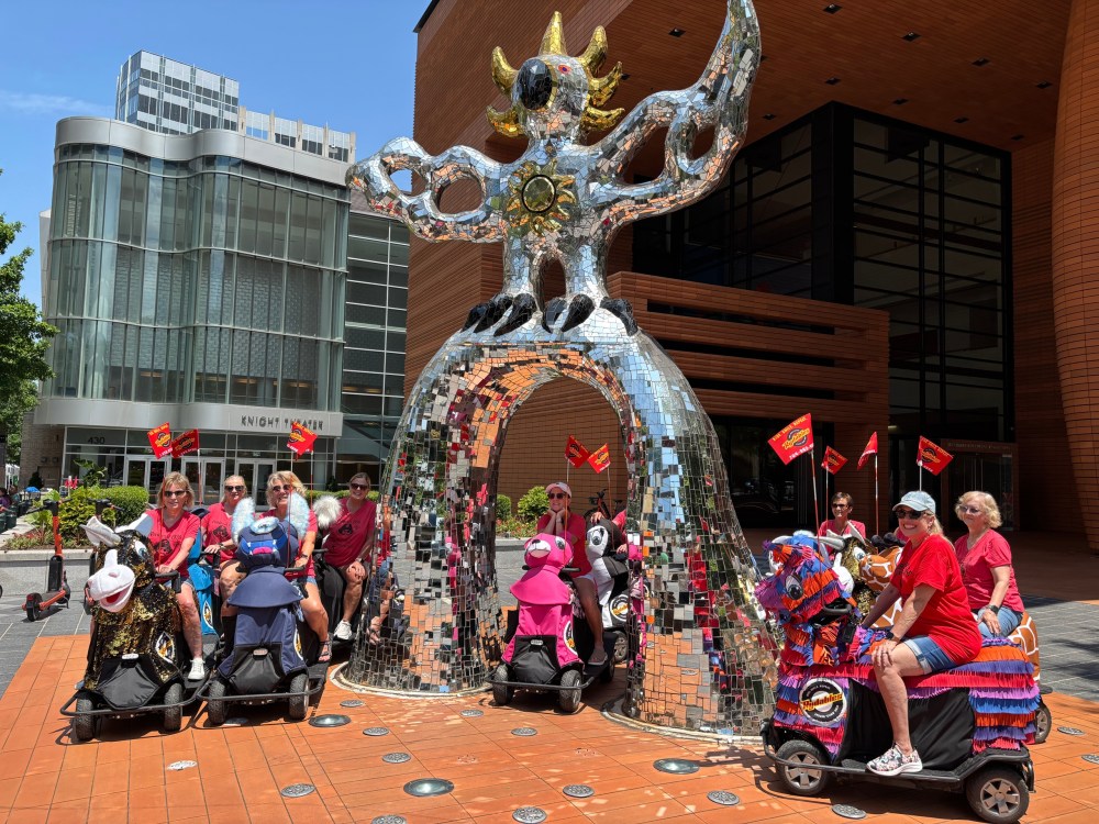 Group of people on mobility scooters with red flags under a reflective sculpture near a theater.