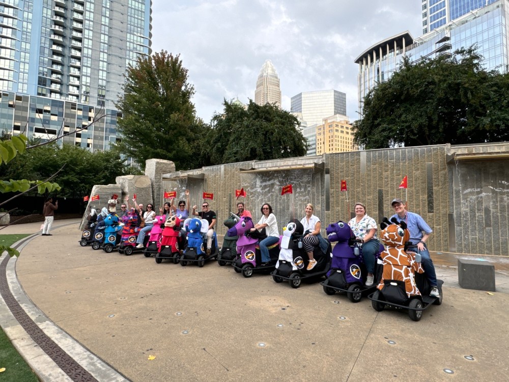 Group of people on motorized animal scooters in a park with city buildings in the background.