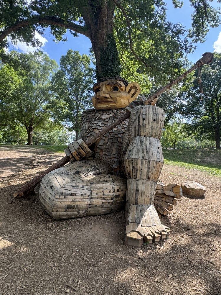 Large wooden troll sculpture sitting under trees.