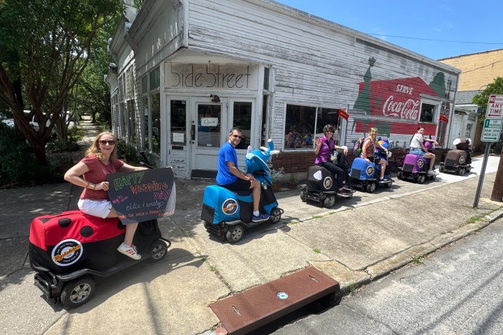 People on scooters in line outside a rustic shop with Coca-Cola mural.