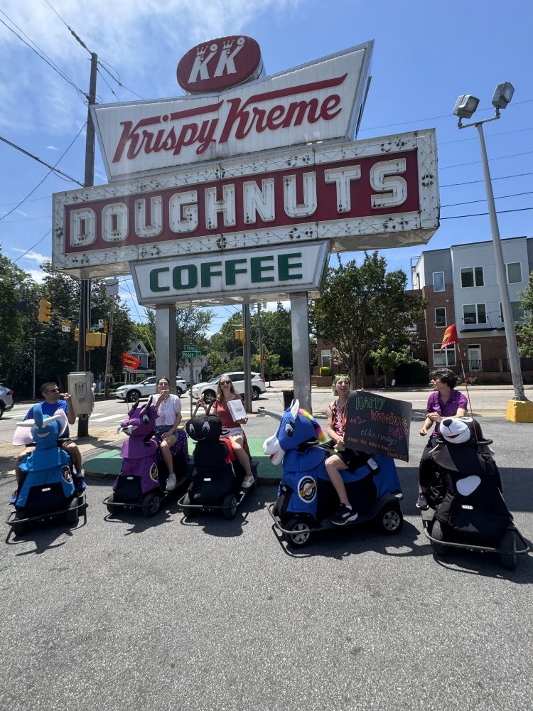Group in costumes and carts under Krispy Kreme Doughnuts sign on a sunny day.