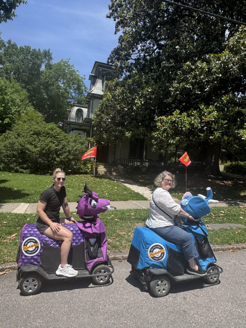 Two people on motorized scooters with colorful covers on a sunny street.