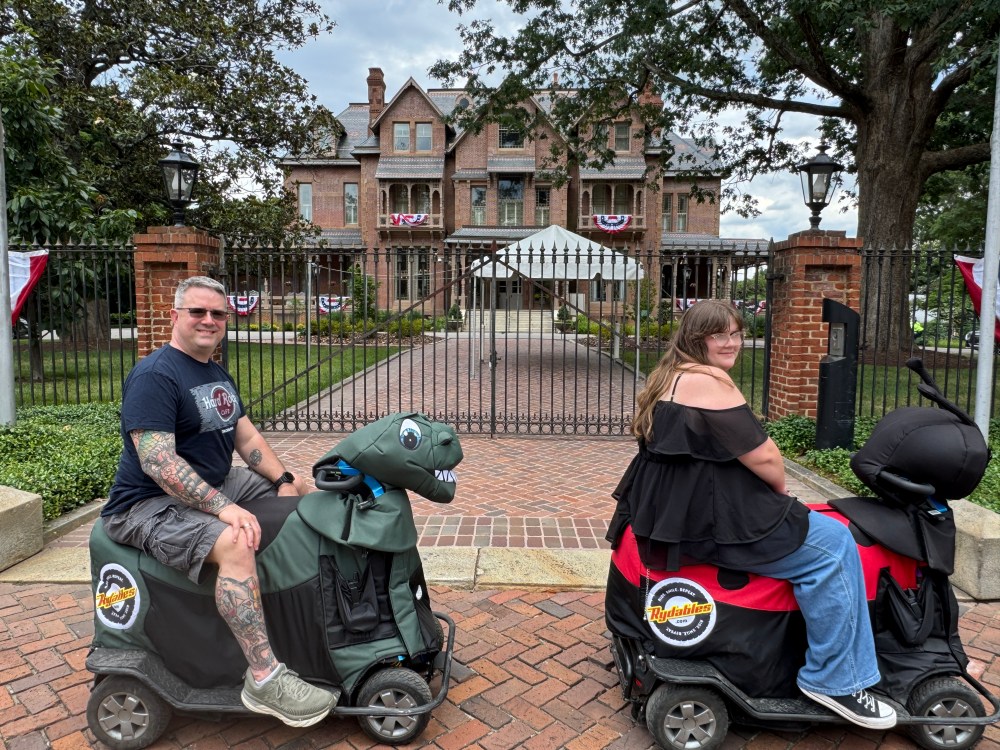 Two people on themed scooters in front of a historic brick mansion.