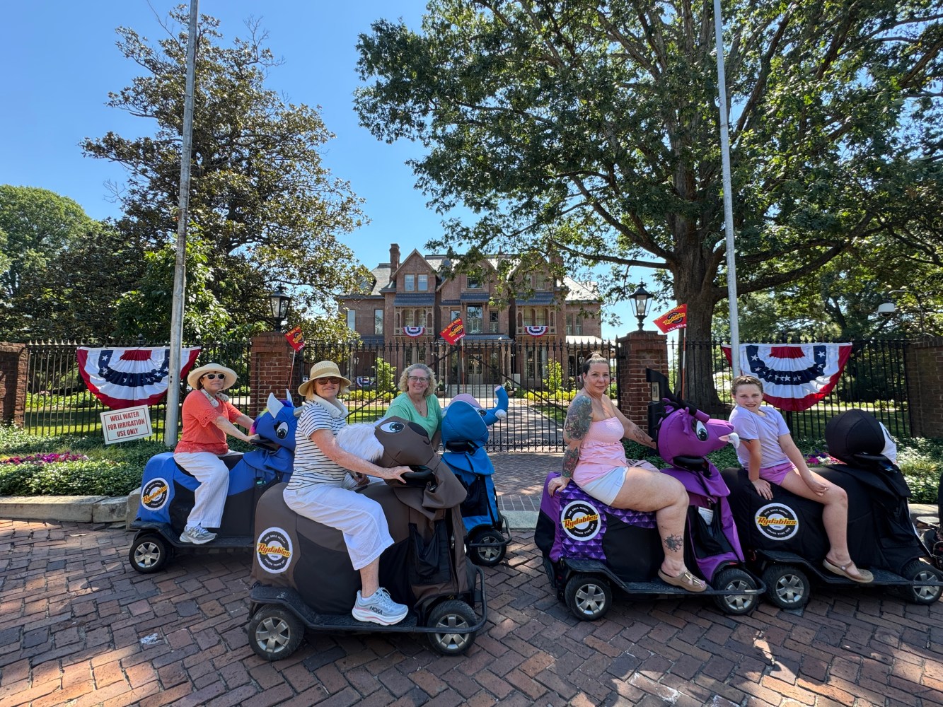 Five people on ride-on scooters in front of a house with patriotic bunting.
