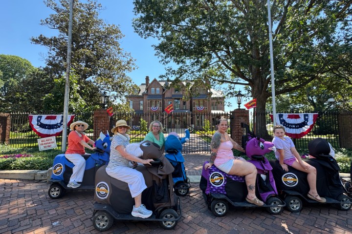 Five people on ride-on scooters in front of a house with patriotic bunting.