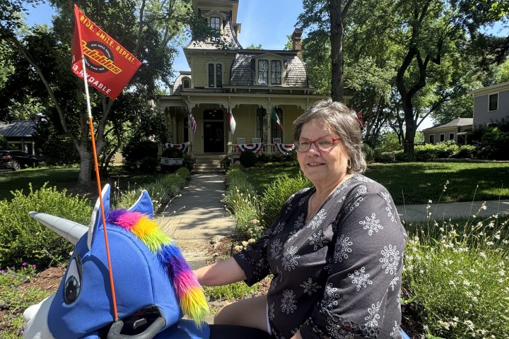 Woman sitting on blue unicorn bike in garden with house in background.