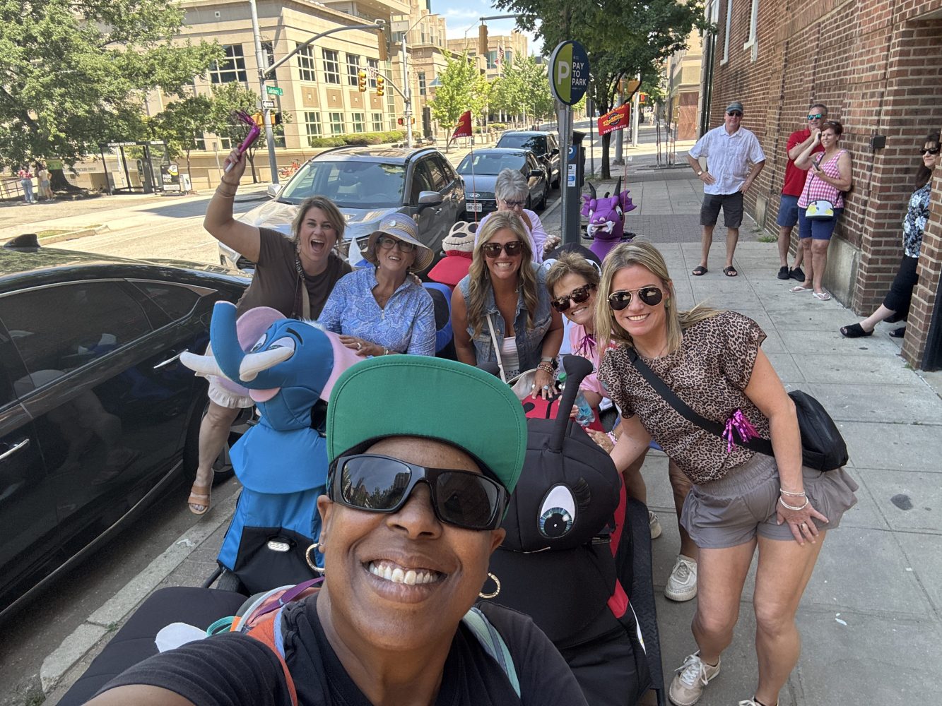Group selfie of people smiling on a city sidewalk with a bright sunny day.